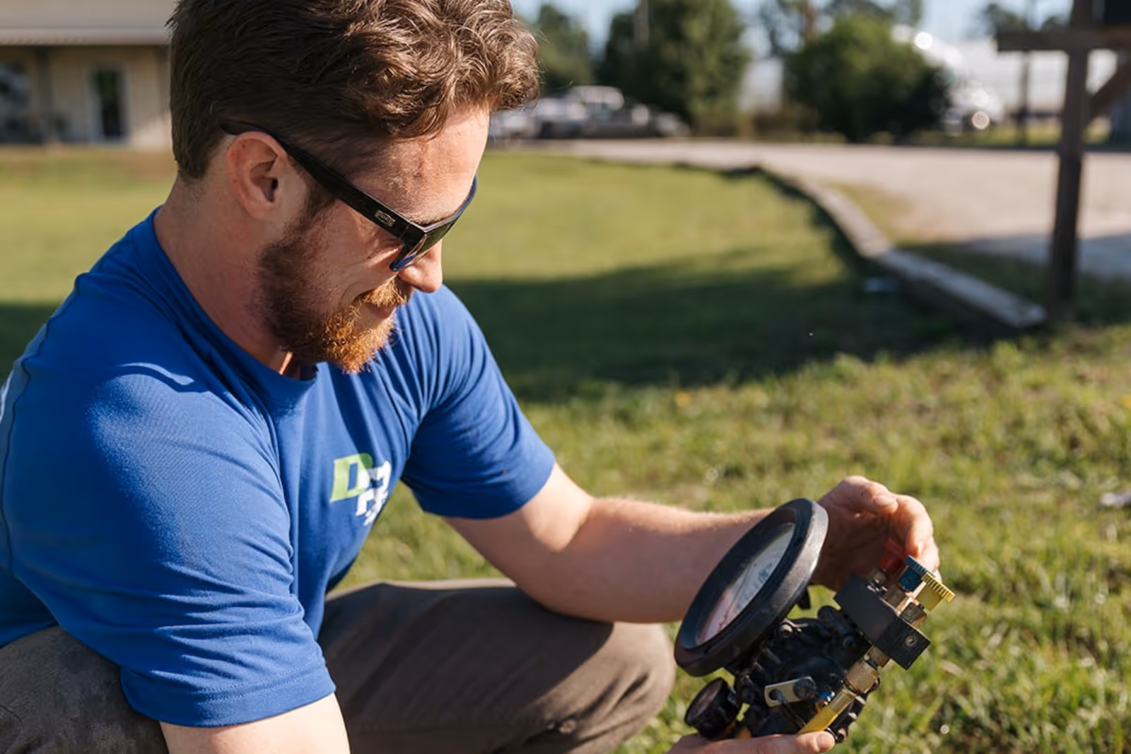 A man wearing a blue, branded Dr Rooter top holds a tool in his hand as he leans forward to closely inspect a drain, examining it with a focused expression.